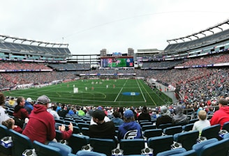 A large sports stadium filled with spectators. The field is green with white markings, and players in red and white uniforms are actively engaged in a game. The stands are packed with people wearing various colored clothing, and some sections of the crowd are more densely populated. Digital screens and advertisements are visible around the stadium, adding to the vibrant atmosphere.