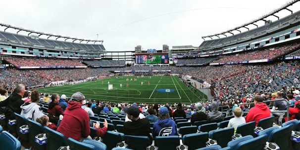 A large sports stadium filled with spectators. The field is green with white markings, and players in red and white uniforms are actively engaged in a game. The stands are packed with people wearing various colored clothing, and some sections of the crowd are more densely populated. Digital screens and advertisements are visible around the stadium, adding to the vibrant atmosphere.