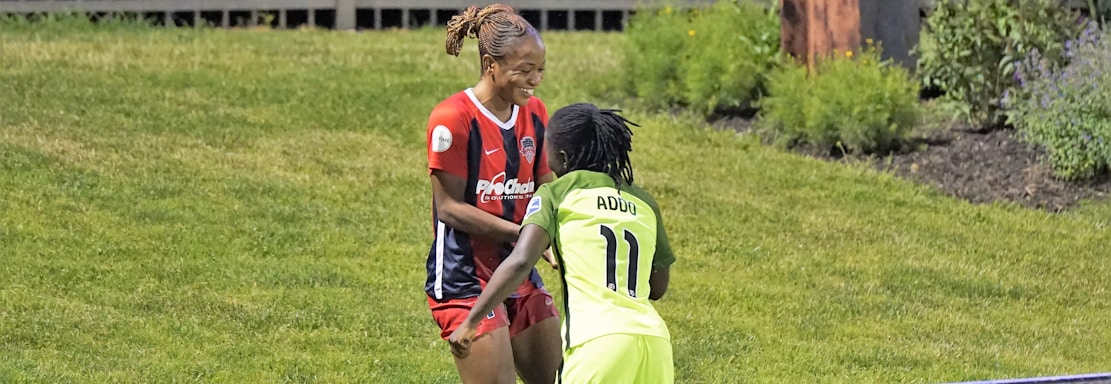 Two individuals are interacting on a grassy field. One is wearing a red and blue sports outfit with 'ProChain' visible, and the other is in a bright yellow uniform with the name 'Addo' and the number 11. They appear to be playing a sport, possibly soccer, as they are near a marked boundary. A fence and some vegetation are visible in the background.