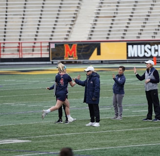 Several people are on a sports field with an empty bleacher in the background. One person is high-fiving a coach, while others stand nearby. They are wearing athletic clothing, hinting at a team sport environment. A sign with the text 'Muscle Milk' is visible in the background.