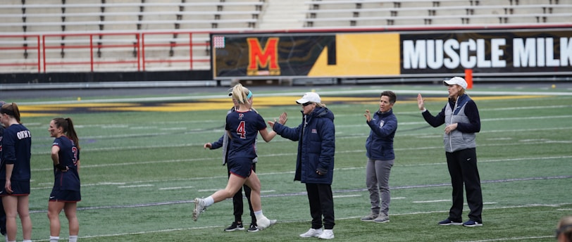 Several people are on a sports field with an empty bleacher in the background. One person is high-fiving a coach, while others stand nearby. They are wearing athletic clothing, hinting at a team sport environment. A sign with the text 'Muscle Milk' is visible in the background.