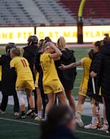 A group of athletes wearing yellow sports uniforms are standing on a sports field. Some are holding lacrosse sticks, and they appear to be interacting casually. Several people are dressed in black coats. In the background, there is a bright sign with text and parts of a stadium are visible.