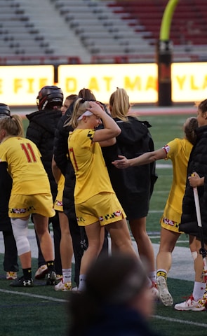 A group of athletes wearing yellow sports uniforms are standing on a sports field. Some are holding lacrosse sticks, and they appear to be interacting casually. Several people are dressed in black coats. In the background, there is a bright sign with text and parts of a stadium are visible.