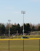 A sports field with a group of people engaging in athletic activities in the background. There is sports equipment like a soccer goal and a scoreboard visible. A chain-link fence runs across the foreground, and a statue or sculpture resembling an eagle is also seen. Trees and tall floodlights are positioned behind the field.