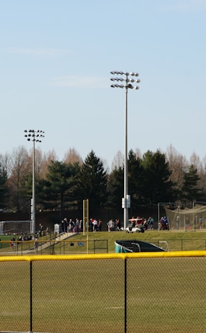 A sports field with a group of people engaging in athletic activities in the background. There is sports equipment like a soccer goal and a scoreboard visible. A chain-link fence runs across the foreground, and a statue or sculpture resembling an eagle is also seen. Trees and tall floodlights are positioned behind the field.
