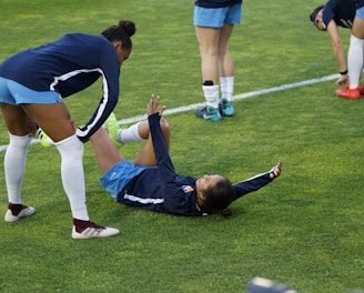 A group of athletes in sportswear are on a green soccer field. Two individuals are interacting closely; one helps the other who is lying on the grass. Nearby, other athletes are stretching.