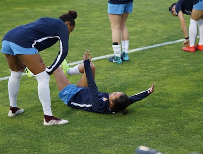 A group of athletes in sportswear are on a green soccer field. Two individuals are interacting closely; one helps the other who is lying on the grass. Nearby, other athletes are stretching.