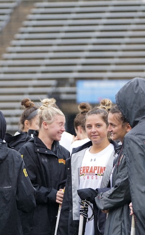 A group of people, likely athletes, are gathered together on a sports field. They are wearing black jackets with some individuals dressed in white sports uniforms underneath. A few people have their hair tied up in buns, and most are engaged in conversation. The background shows tiered stadium seats, suggesting the setting is an outdoor sports venue.