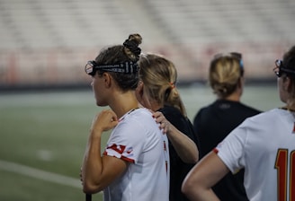Several people wearing sports attire and protective eyewear are gathered on a sports field. They appear to be engaged in a discussion or briefing, possibly during a break from a game or practice. The background shows a blurred stadium seating, indicating an outdoor setting.