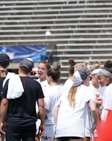 A group of athletes in white shirts with black and red accents are gathered together, engaged in conversation and smiling. They appear to be on a sports field with empty bleachers in the background. The setting is outdoors, and it seems sunny. Some individuals are wearing caps or headbands.