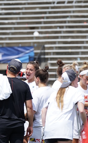 A group of athletes in white shirts with black and red accents are gathered together, engaged in conversation and smiling. They appear to be on a sports field with empty bleachers in the background. The setting is outdoors, and it seems sunny. Some individuals are wearing caps or headbands.