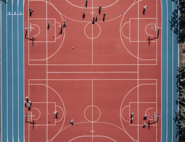 An aerial view of a large, vibrant sports complex featuring a red basketball court with multiple hoops and a surrounding blue running track. Several people are scattered across the area, engaged in various activities.