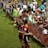 The image features a lively gathering of young soccer players and fans next to a field. Several female athletes in red and blue uniforms interact with an enthusiastic crowd standing behind a barrier. Some people in the crowd are holding objects, like pens and paper, possibly for autographs.