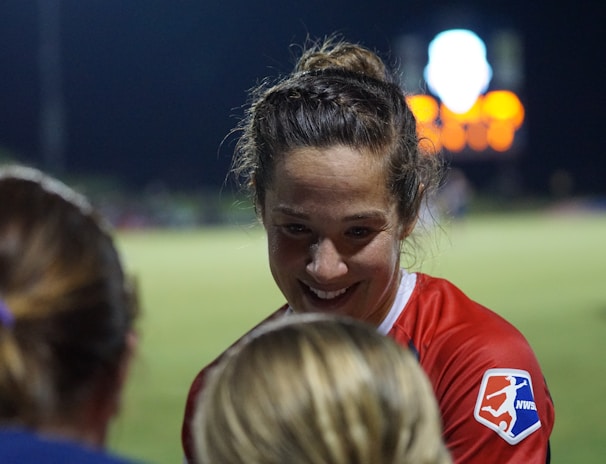 A female athlete in a red sports jersey with the NWSL logo energetically interacts with others on a sports field. The backdrop consists of a darkened night sky and a blurred scoreboard or advertisement display, with soft, colorful lights.
