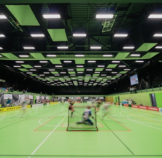 A sports hall with bright green flooring and ceiling panels. There is a game in progress with players moving quickly, suggesting a fast-paced sport like floorball or indoor hockey. Advertising banners line the walls, and a scoreboard is visible in the background.