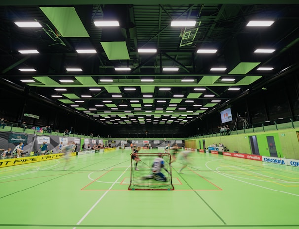 A sports hall with bright green flooring and ceiling panels. There is a game in progress with players moving quickly, suggesting a fast-paced sport like floorball or indoor hockey. Advertising banners line the walls, and a scoreboard is visible in the background.