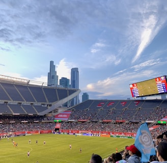 A sports stadium filled with spectators watching a soccer match. Players are on the field in action, and a large scoreboard displays information about the game. The stadium has blue seating and is surrounded by tall city buildings visible in the background against a partly cloudy sky. Fans are dressed casually, some waving flags and wearing team colors.