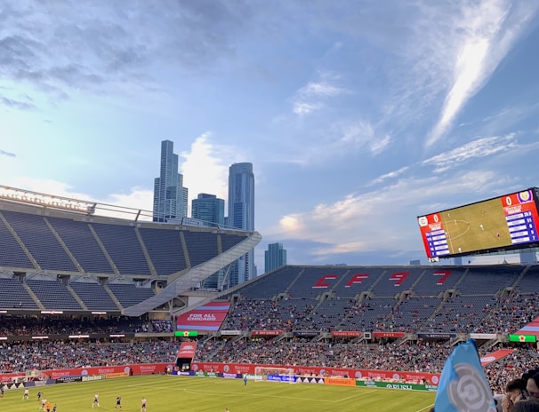 A sports stadium filled with spectators watching a soccer match. Players are on the field in action, and a large scoreboard displays information about the game. The stadium has blue seating and is surrounded by tall city buildings visible in the background against a partly cloudy sky. Fans are dressed casually, some waving flags and wearing team colors.