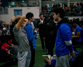 People engage in conversation and activities in an indoor sports facility. A woman in gray athletic wear stretches her arms above her head while talking to a man in a blue jacket. Other people are seen interacting in the background, some are seated on the ground, and the atmosphere is lively with many individuals gathered.